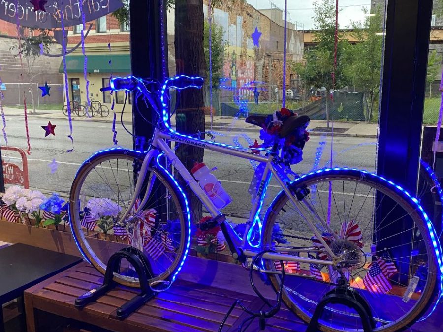 Bike with blue lights, flowers, and flags in window of Earth Rider bike shop celebrating July 4th and cycling freedom