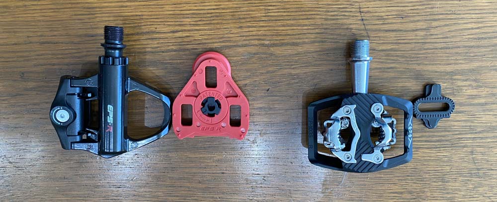 Clipless road pedal with a matching three-hole cleat next to a multipurpose pedal with a SPD style two-hole cleat.