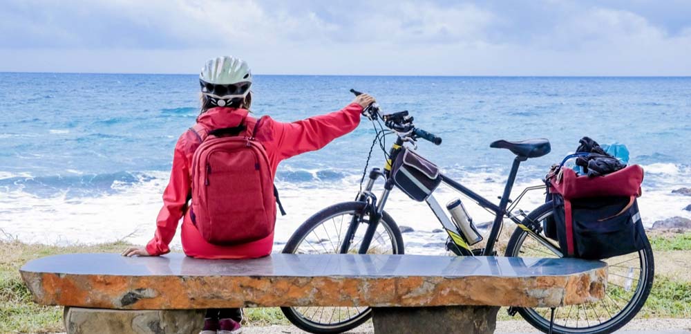 Cyclist wearing a jacket and backpack holding a bike gazing onto the ocean.