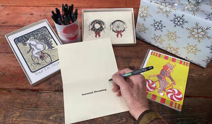 Person writing a message in a greeting card on a desk surrounded by artist designed holiday cards